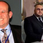 Two men in business suits: left a smiling man with glasses and a blue lanyard; right a seated man in a dark suit at a desk.