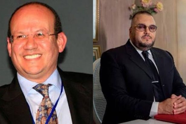 Two men in business suits: left a smiling man with glasses and a blue lanyard; right a seated man in a dark suit at a desk.