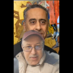 Two men posing for a selfie in front of a colorful mural; the foreground man wears glasses, a pink cap, and a beige jacket.