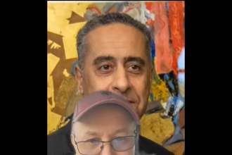 Two men posing for a selfie in front of a colorful mural; the foreground man wears glasses, a pink cap, and a beige jacket.