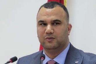 Man in a blue suit and pink tie speaking at a podium with two microphones, flag visible in the background.