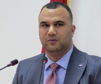 Man in a blue suit and pink tie speaking at a podium with two microphones, flag visible in the background.