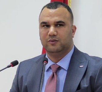 Man in a blue suit and pink tie speaking at a podium with two microphones, flag visible in the background.