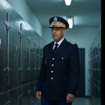 Officer in navy dress uniform with peaked cap standing in a locker-room hallway lined with metal lockers.