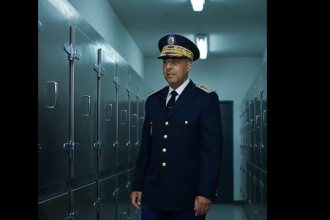 Officer in navy dress uniform with peaked cap standing in a locker-room hallway lined with metal lockers.