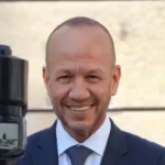 Smiling man in a navy suit and tie posing for a photo outdoors, with camera gear visible on the left side.