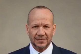 Man wearing a dark suit and tie, facing the camera with a neutral expression against a plain background.