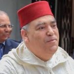 Man wearing a red fez and a light-colored traditional robe, outdoors with another blurred person in the background.