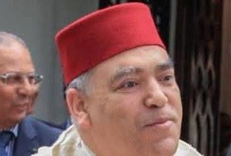 Man wearing a red fez and a light-colored traditional robe, outdoors with another blurred person in the background.