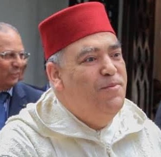 Man wearing a red fez and a light-colored traditional robe, outdoors with another blurred person in the background.