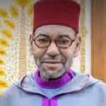 Portrait of a smiling man wearing a red fez and purple robe, with round glasses, indoors.