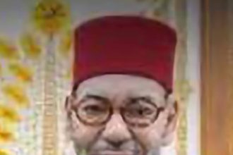Portrait of a smiling man wearing a red fez and purple robe, with round glasses, indoors.