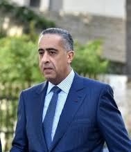Man in a blue suit and light blue tie walking outdoors, with greenery and a stone building in the background.