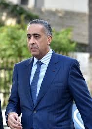 Man in a blue suit and light blue tie walking outdoors, with greenery and a stone building in the background.