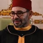 Man with glasses in a red fez and black robe with yellow trim, seated indoors.