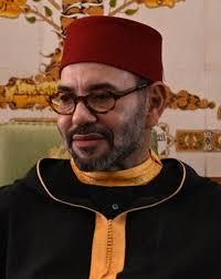 Man with glasses in a red fez and black robe with yellow trim, seated indoors.
