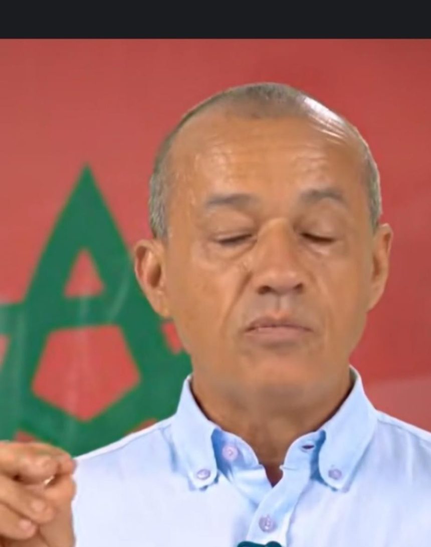 Older man with short gray hair and a light blue button-down shirt, eyes closed, in front of a red backdrop.